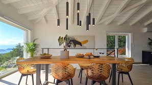 Un comedor con una mesa de madera, seis sillas, lámparas colgantes, una planta en maceta y un cuadro en la pared.