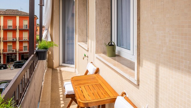 A balcony with a wooden table and chairs, a potted plant, and a window with a curtain. - 605A Residenza Gigli (Garda)