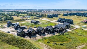 Un resort moderno y ecológico con varias cabañas y un edificio central, rodeado de campos abiertos y un pueblo al fondo.