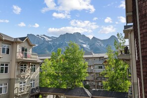 A multi-story building with balconies, a view of snow-capped mountains, and green trees in the foreground. - Spacious High-End Air Conditioned, 2 Bedroom 2 Bathroom Condo In Canmore/Banff. (Canmore)