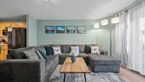 Ein modernes Wohnzimmer mit einer Eckcouch, einem Couchtisch aus Holz und einer Küchenzeile mit Kühlschrank und Schränken.