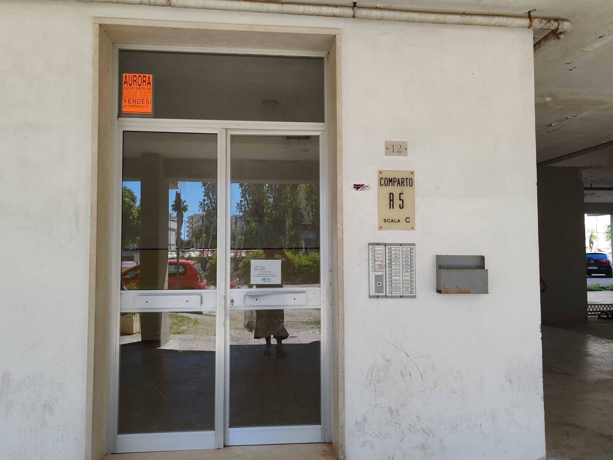 A building entrance with a glass door, a sign for 'AURORA' indicating a real estate agency, and a 'COMPARTO A5' sign with a 'SCALA C' label.