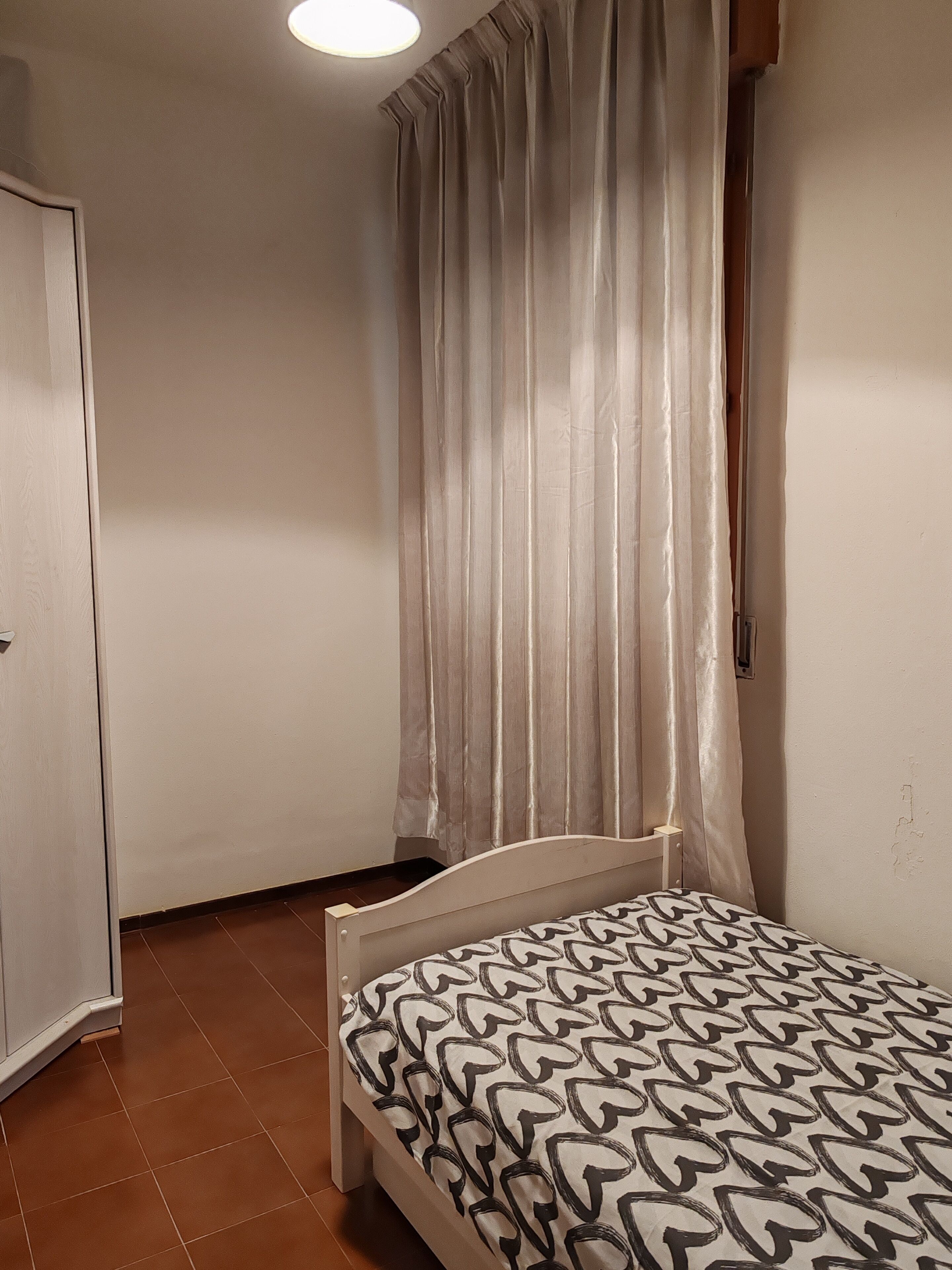 Ein kleiner, sauberer Raum mit einem Einzelbett, einer Tür und einem Fenster mit Vorhängen.