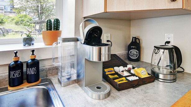 Eine Küche mit Kaffeemaschine, Wasserkocher und Kaffeezubereiter. Auf der Arbeitsplatte stehen zwei Flaschen mit den Aufschriften 'Dishes' und 'Hands'.