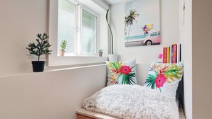 Ein Schlafzimmer mit Fenster, einer kleinen Pflanze, einem Gemälde und einem Bücherregal.