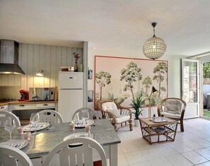 A modern kitchen with white cabinets, a dining area with white chairs, a wicker coffee table, and a large window with a view of greenery.