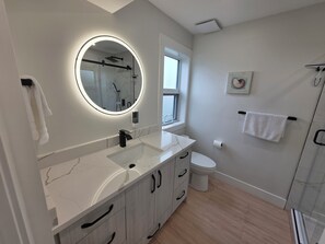 Un baño moderno con lavamanos de mármol blanco, un espejo redondo con iluminación ambiental, inodoro y cabina de ducha de vidrio.