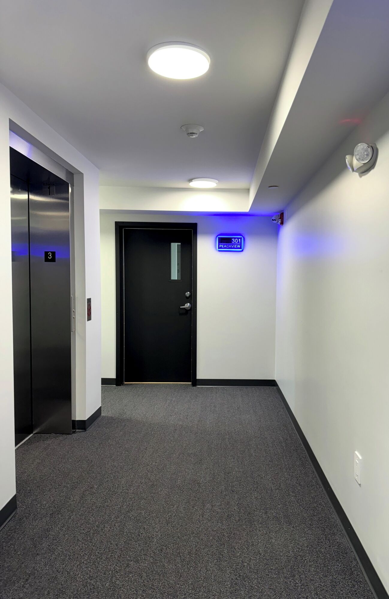 Un pasillo con una puerta negra, un letrero iluminado en azul y un ascensor.