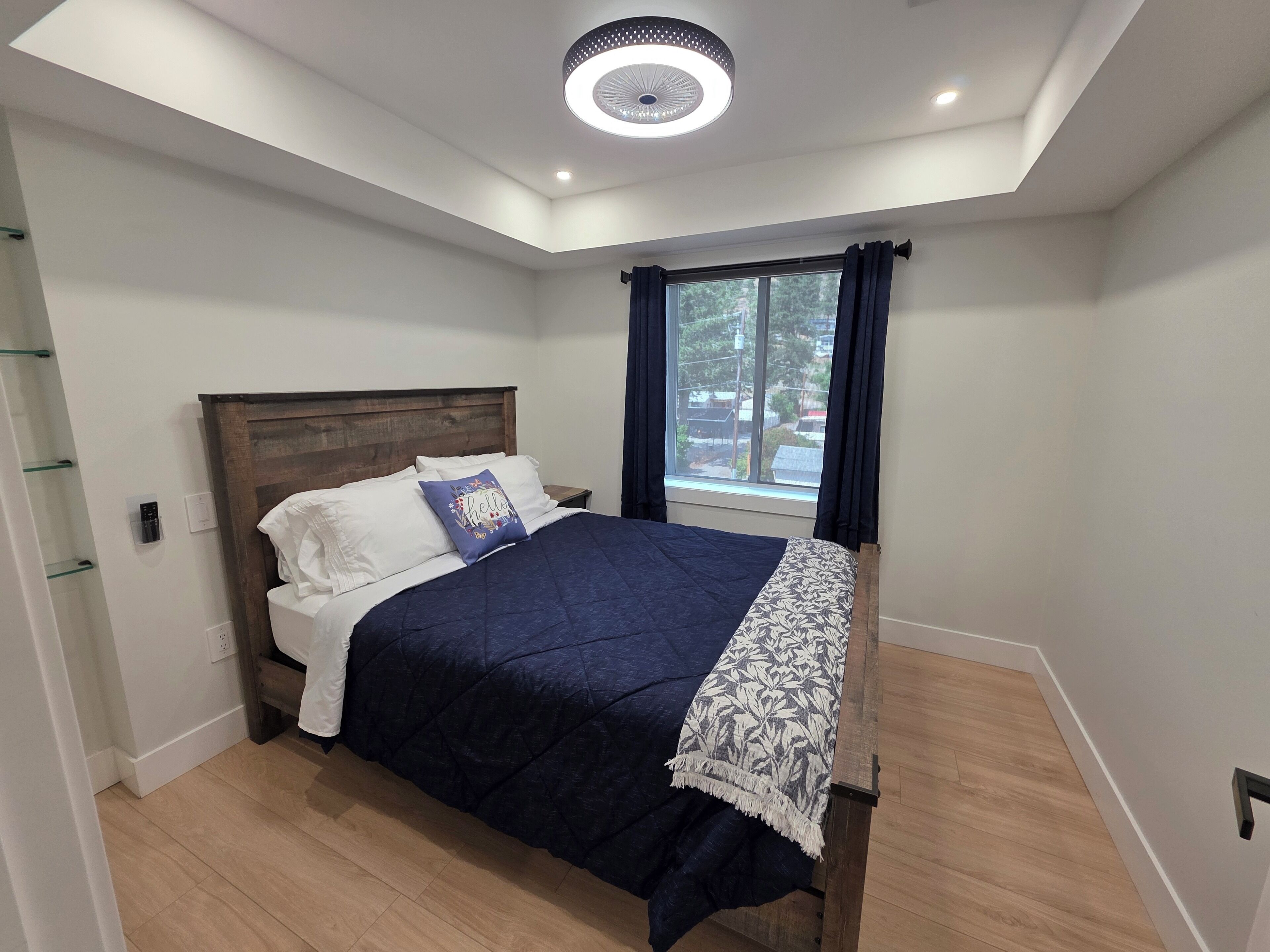 Une chambre moderne avec une tête de lit en bois, un grand lit recouvert d’une couverture bleu foncé et agrémenté d’un plaid à motifs, des oreillers blancs et une fenêtre avec des rideaux foncés.