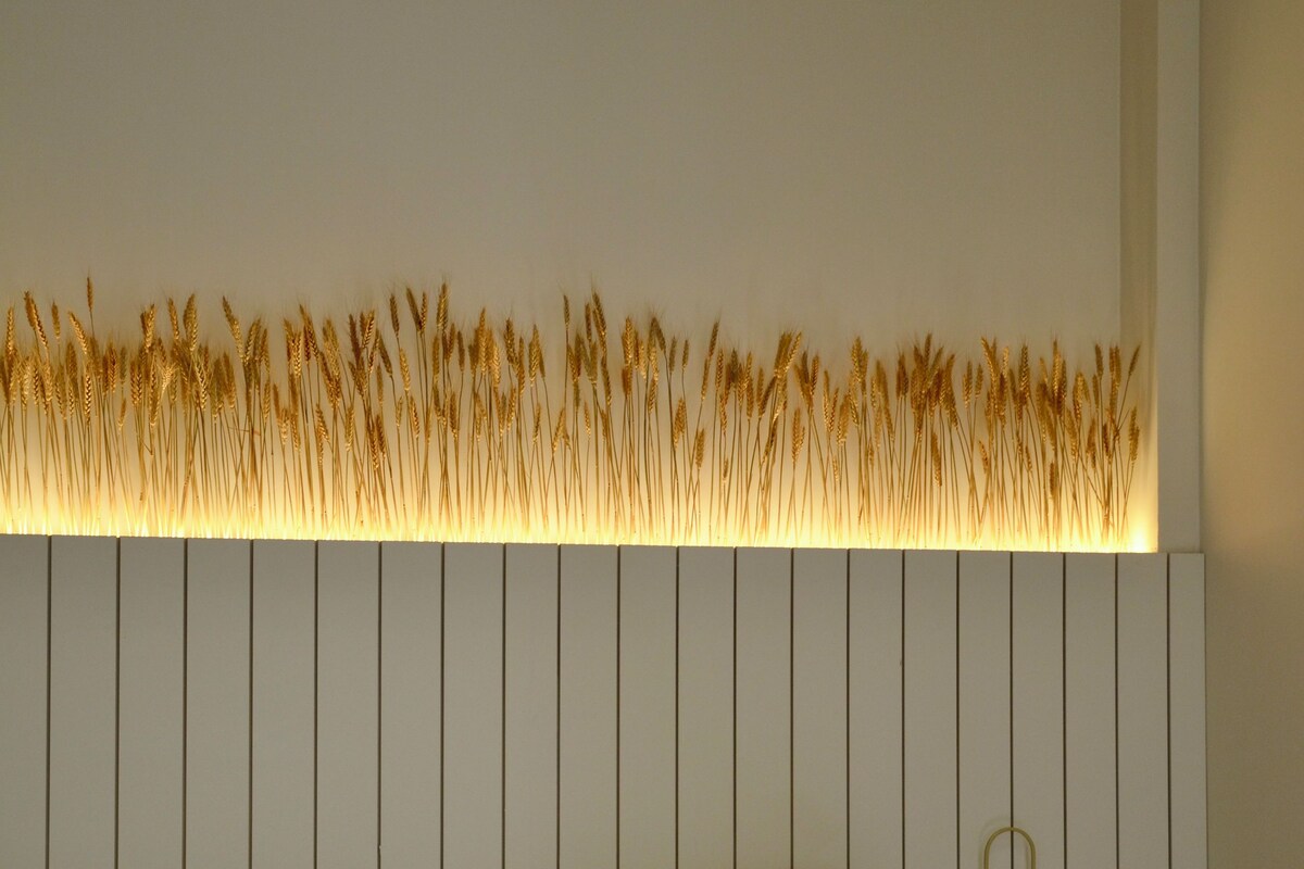 A hotel room with a bed, white linens, a wall-mounted light fixture with a golden wheat design, and a wall socket.