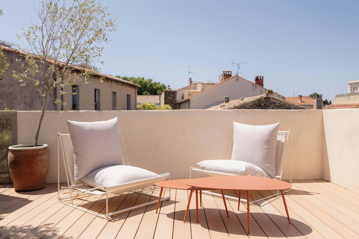 A terrace with two white cushioned chairs and a small table, a potted plant, and a view of buildings in the background.