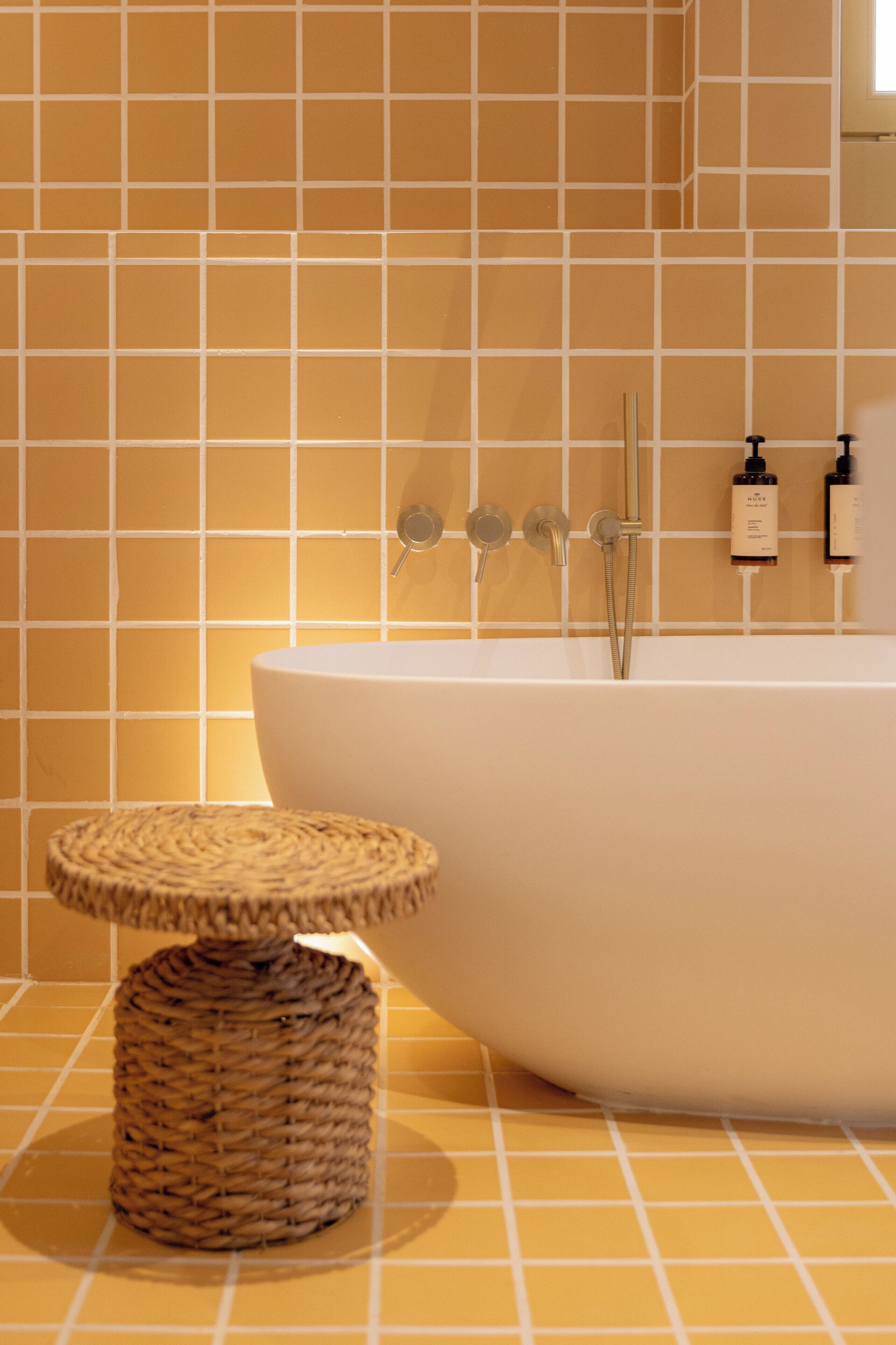 Ein Badezimmer mit einer großen Badewanne, einem kleinen runden Korbstuhl und einer gefliesten Wand mit Rasteroptik.