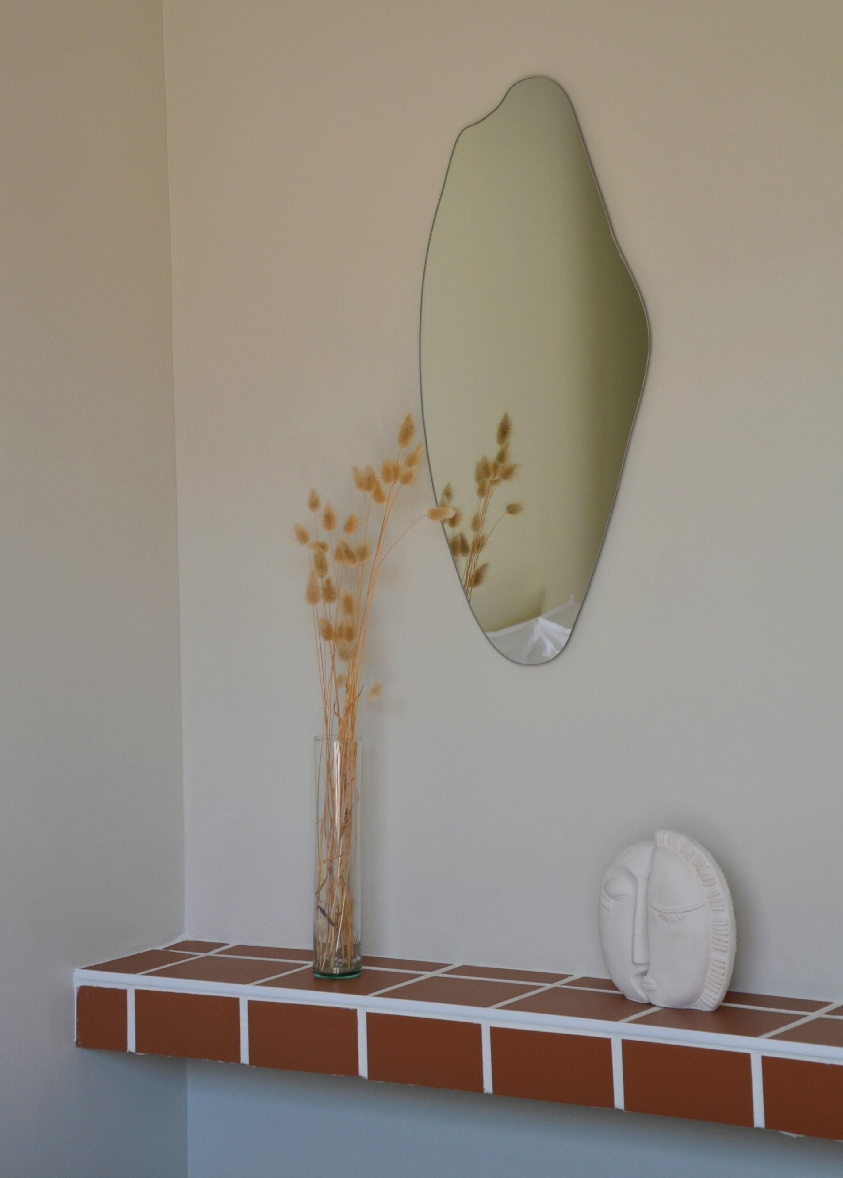 Een spiegel aan de muur, een vaas met gedroogde planten en een decoratief gezichtsbeeld op een plank.