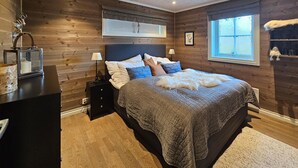 Een slaapkamer met een houten wand, een groot bed met veel kussens, een nachtkastje, een lamp en een raam met uitzicht.