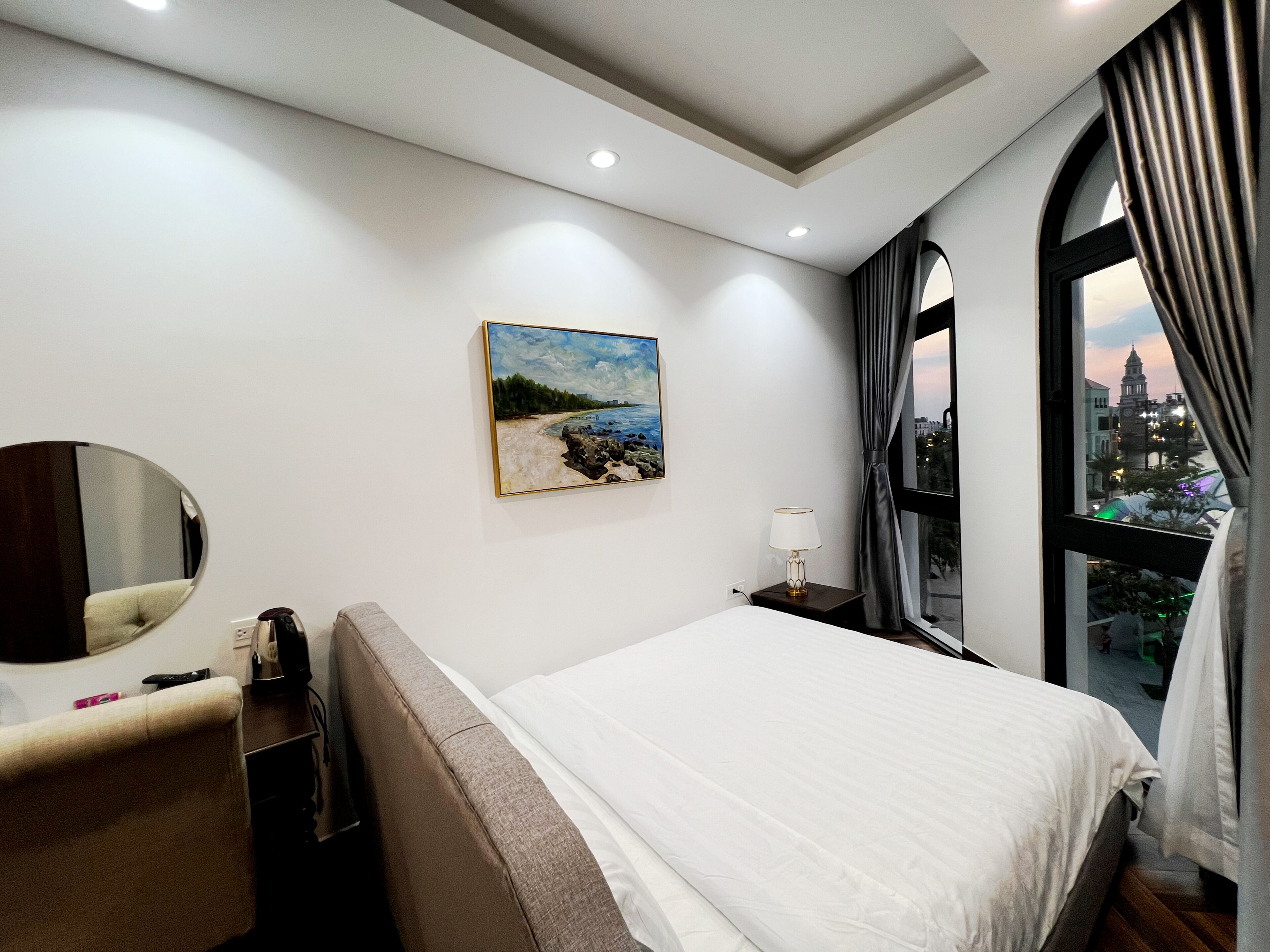 A hotel room with a bed, a bedside table with a lamp, a painting on the wall, a window with curtains, and a view of a cityscape.