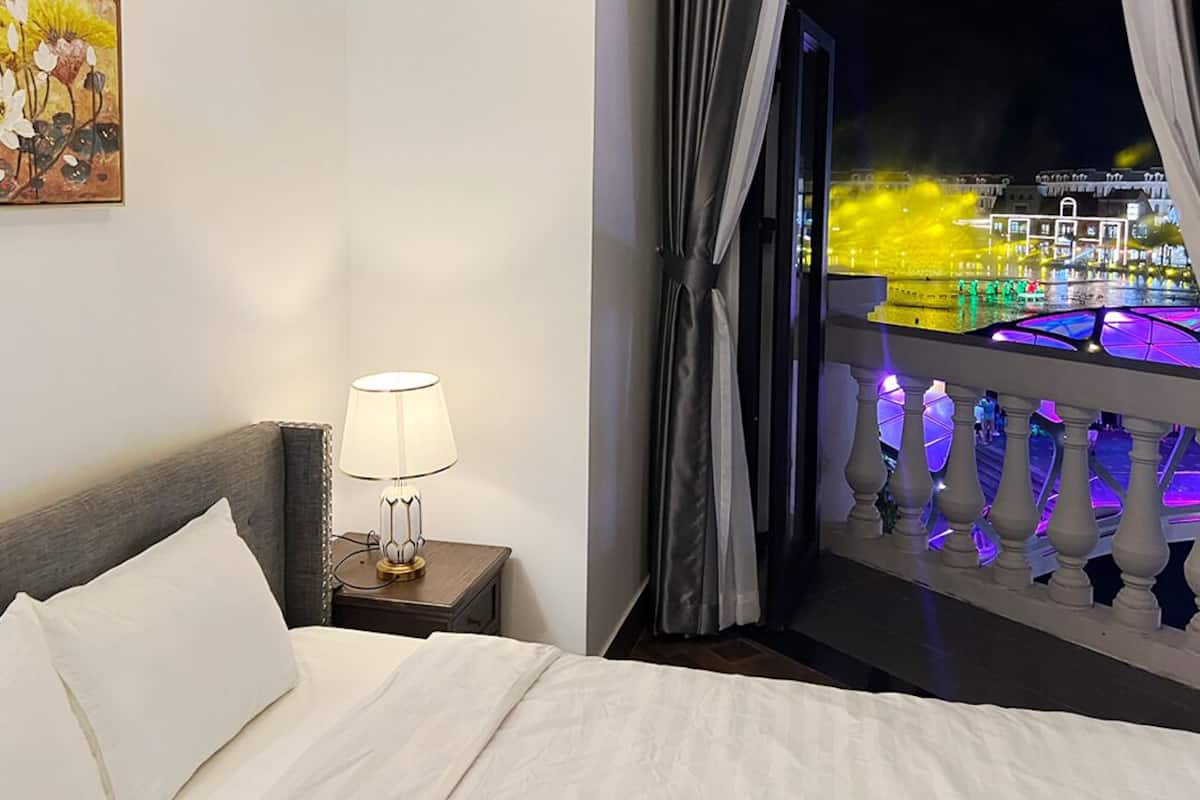 A hotel room with a bed, a nightstand with a lamp, and a view of a lit-up building through a balcony with curtains.