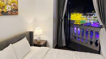 Habitación de hotel con cama, mesita de noche con lámpara y vista a un edificio iluminado a través de un balcón con cortinas.