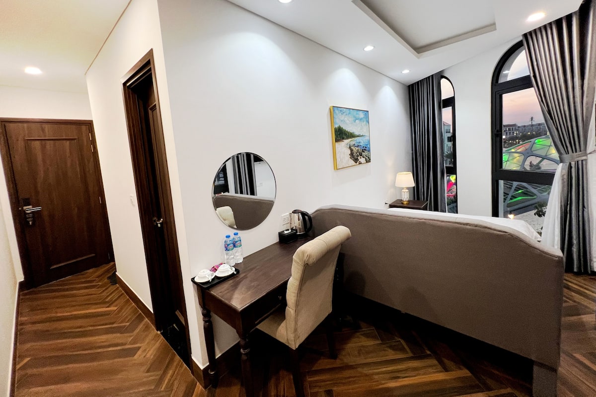 A hotel room with a bed, a desk, a chair, a mirror, a painting, and a view of a cityscape through a window.