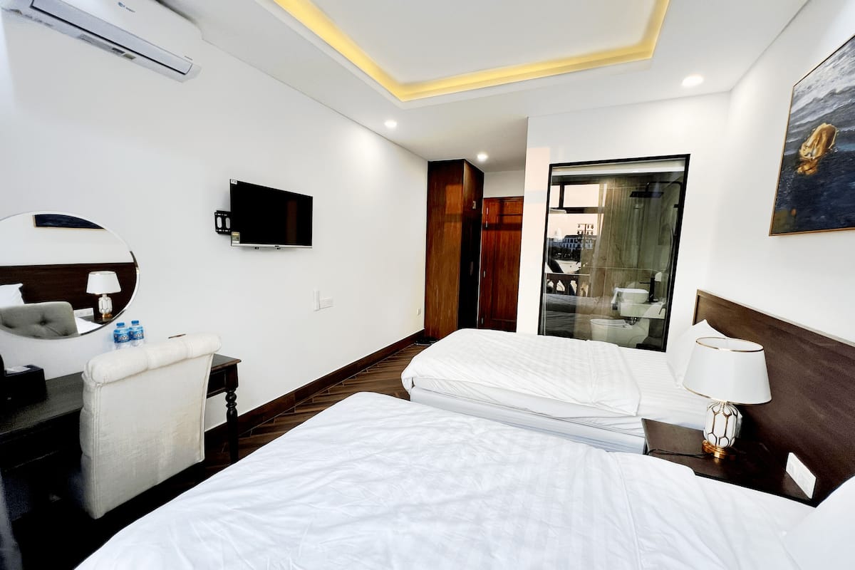 A hotel room with two beds, a TV, a mirror, a desk, and a bathroom visible through a glass door.