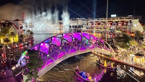 Een helder verlichte voetgangersbrug met een glazen constructie, omgeven door een levendig watergebied met boten en mensen.