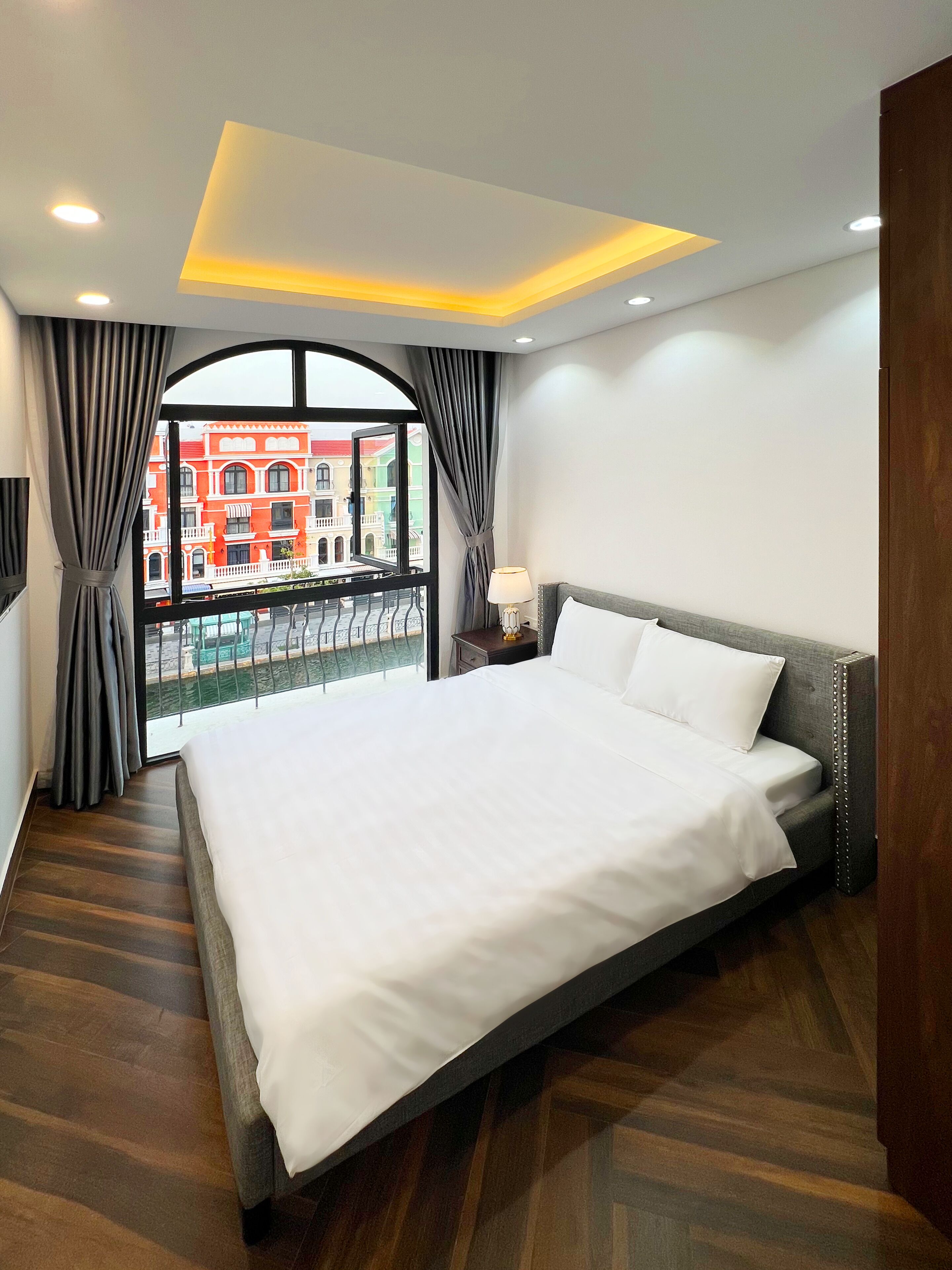 Habitación de hotel moderna con una cama grande, suelo de madera, un balcón con vistas a edificios coloridos e iluminación empotrada en el techo.