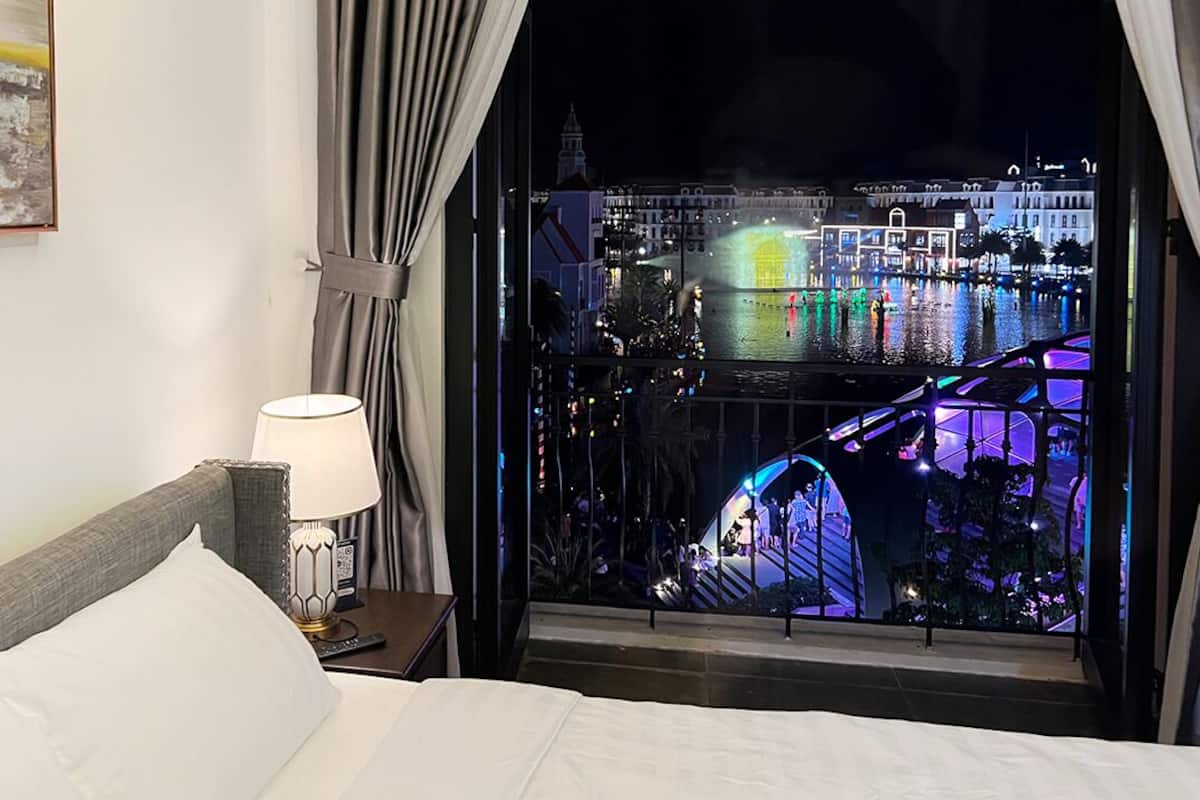A hotel room with a large window overlooking a cityscape at night, featuring illuminated buildings and a water body.