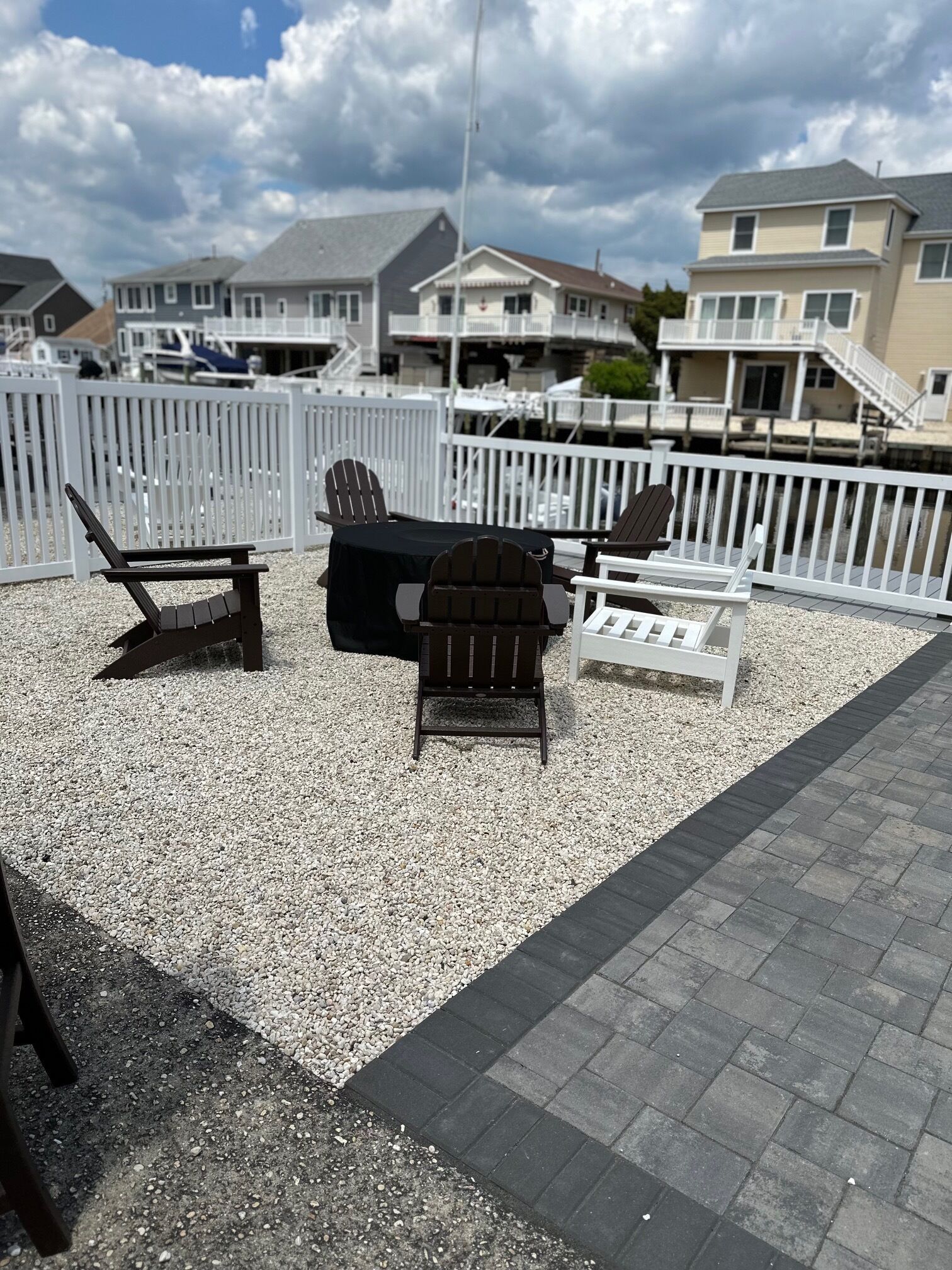 An outdoor patio area with Adirondack chairs and a grill, surrounded by a white fence and a view of houses in the background.