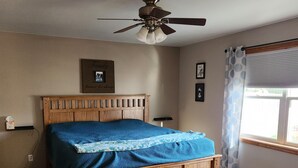 Un dormitorio con una cama de madera, un ventilador de techo, una ventana con cortinas y un cuadro colgado en la pared.