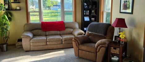 A living room with a beige sectional sofa, a recliner, a ceiling fan, a red lamp, and a view of a green landscape through large windows.