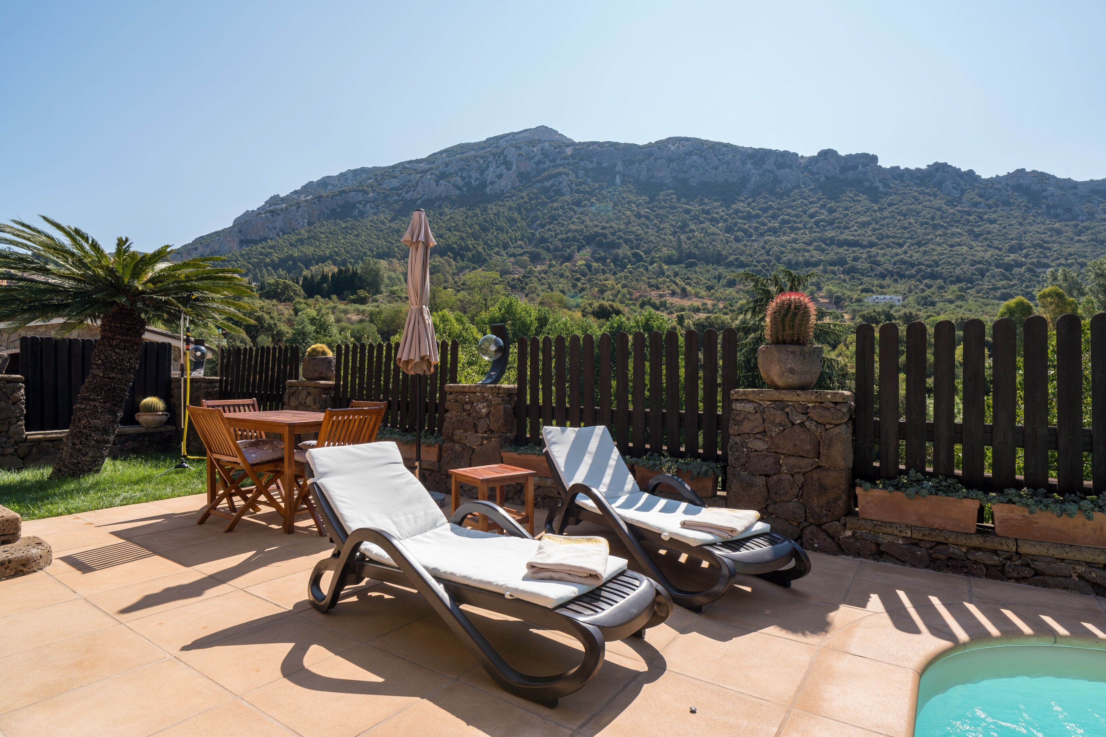 A patio with lounge chairs, a table, and a pool, surrounded by a wooden fence and a mountain backdrop.