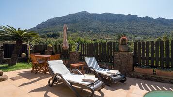 A patio with lounge chairs, a table, and a pool, surrounded by a wooden fence and a mountain backdrop.