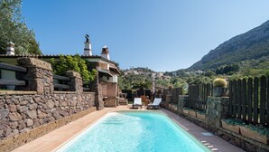 Una piscina con un muro de piedra y una valla de madera, rodeada de vegetación y una montaña al fondo.