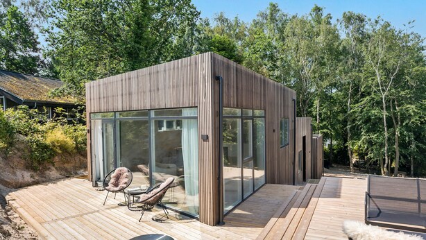 A modern wooden cabin with large glass windows, a deck with lounge chairs, and a view of the surrounding forest.