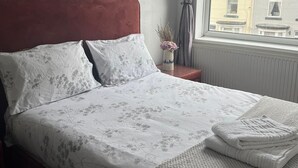Un lit bien fait, avec une literie à motifs floraux, une tête de lit rouge et une vue sur les bâtiments par une fenêtre ornée de rideaux.