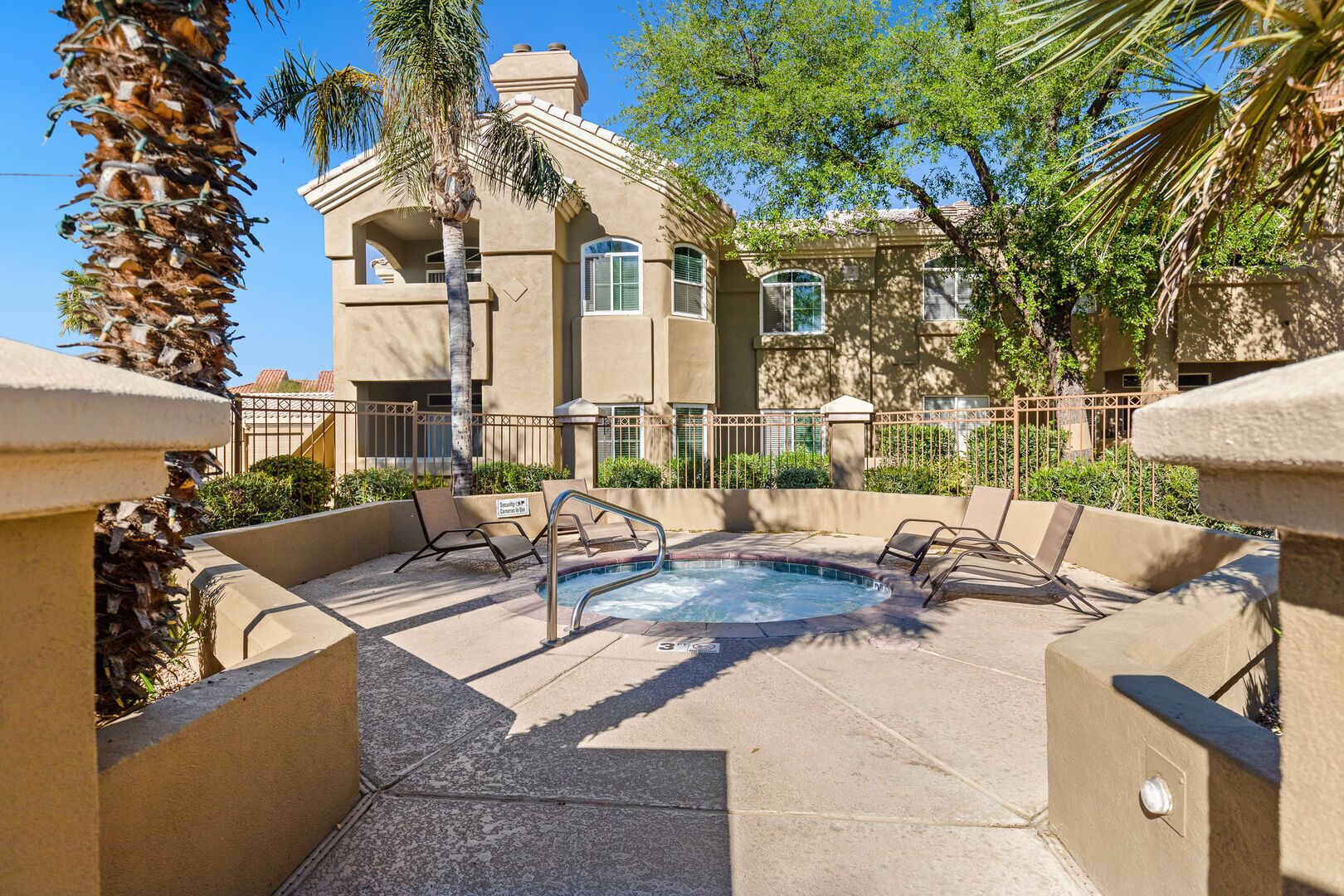 A hot tub surrounded by lounge chairs, with a pool in the background and a building with arched windows.