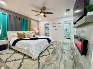 Une chambre moderne avec un lit, un ventilateur de plafond, un téléviseur fixé au mur et une salle de bain visible à travers une porte ouverte.