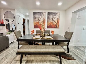 Un coin repas avec une table en bois foncé, dressée pour quatre personnes, entourée de chaises grises, d’un banc et de deux tableaux abstraits accrochés au mur.