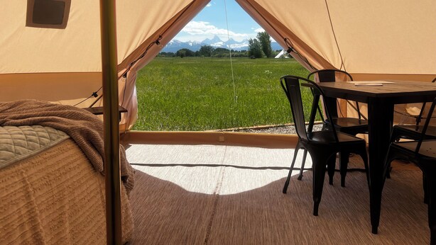 A tent with a table and chairs inside, a bed, and a view of a green field and mountains. - Meadow View Tent Glamping (Tetonia)
