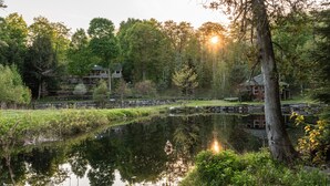 A serene lakeside setting with a stone bridge, a wooden cabin, and a stone house. - Tranquil Oasis in Trent Lakes w/ Private Ponds, Fire Pit, Game Room & Hot Tub (Bobcaygeon)