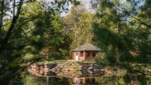 A cabin with a stone foundation, surrounded by a pond and dense forest. - Tranquil Oasis in Trent Lakes w/ Private Ponds, Fire Pit, Game Room & Hot Tub (Bobcaygeon)