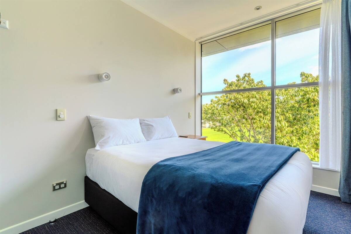 Une chambre d’hôtel avec une grande fenêtre, un lit recouvert de draps blancs et une couverture bleue.