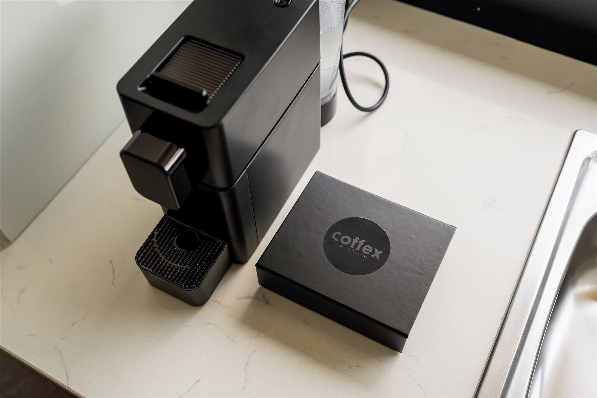 A black coffee machine and a black box with the word "coffex" on a white marble countertop.