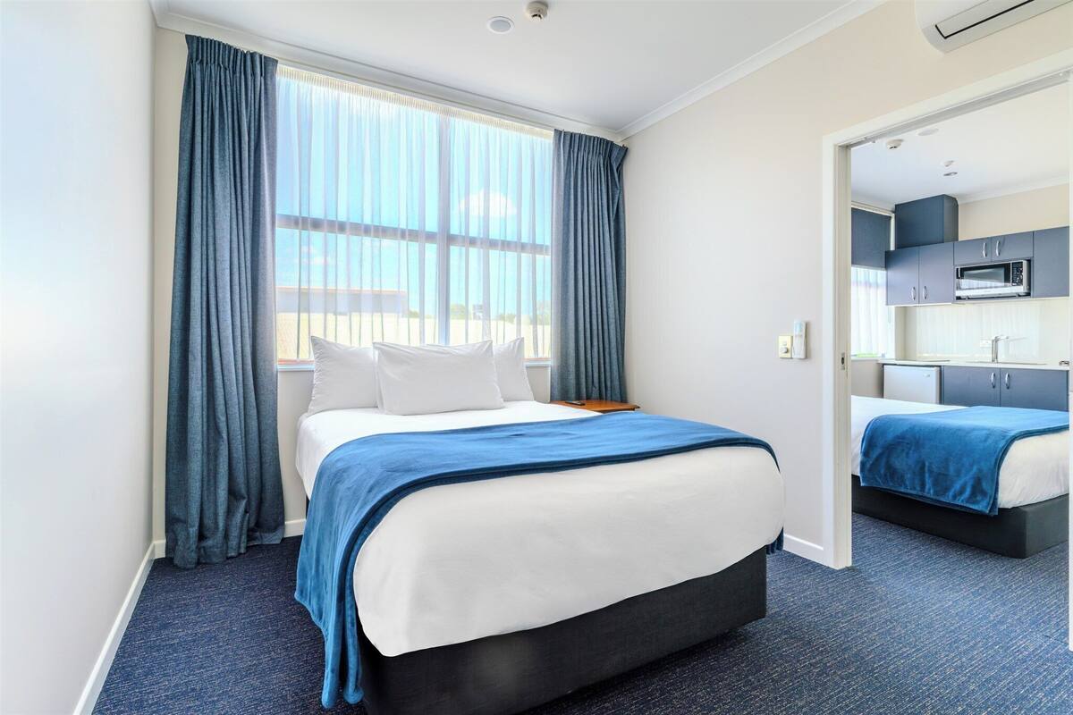 Une chambre d’hôtel comprenant un lit, un petit bureau, une fenêtre avec des rideaux et un coin cuisine.