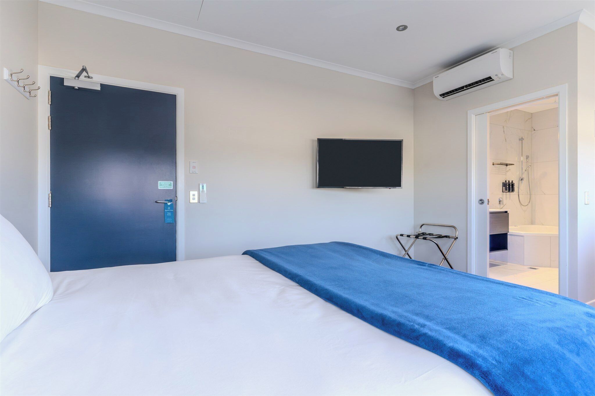 Une chambre d’hôtel avec un lit, un téléviseur fixé au mur, un appareil de climatisation et une salle de bain visible à travers une porte ouverte.