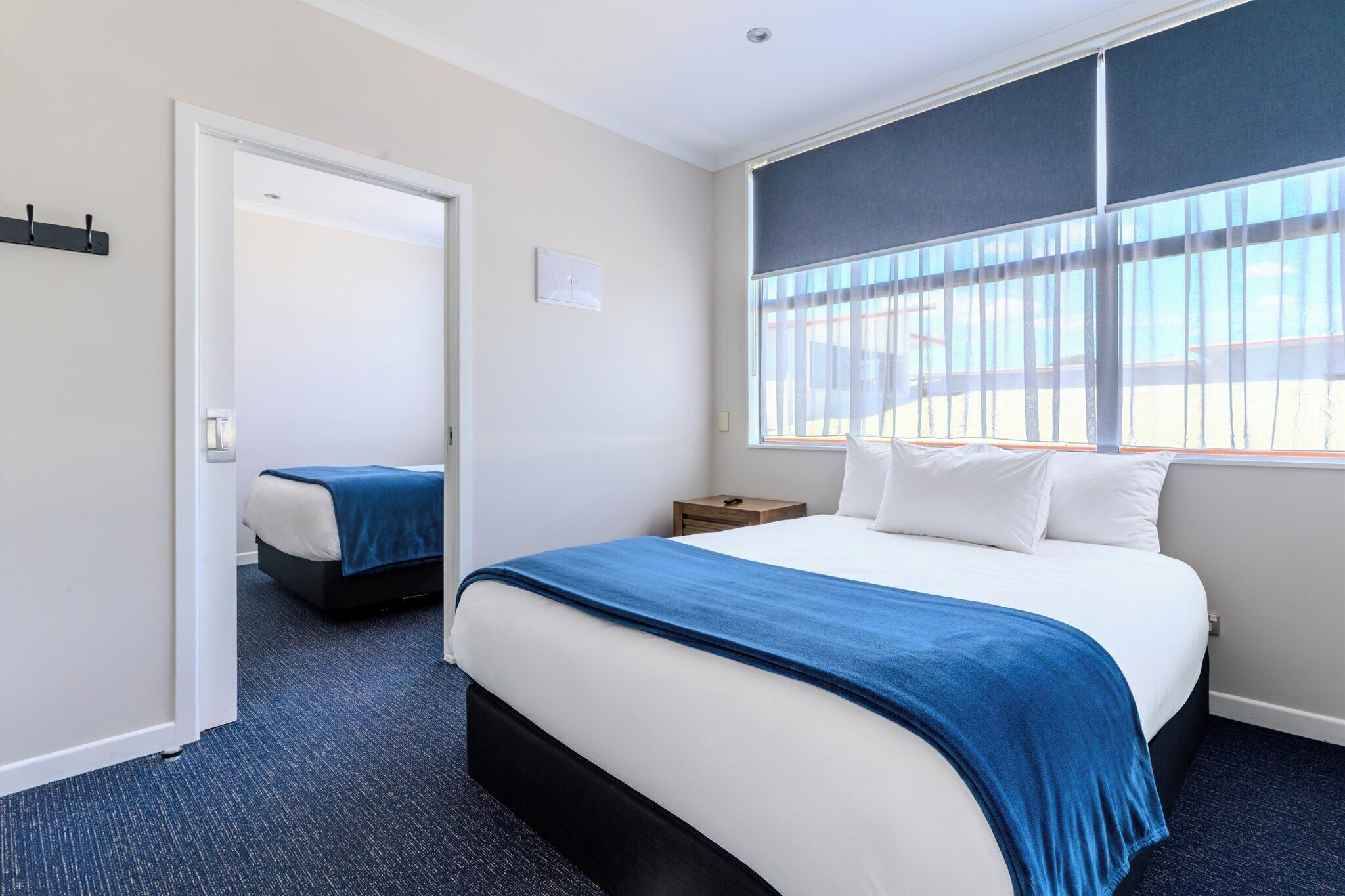 Una habitación de hotel con una cama grande, una mesita de noche, una ventana con persianas y una puerta que conduce a otra habitación.