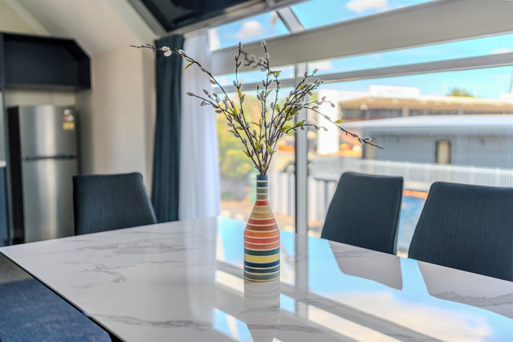 Ein moderner Essbereich mit einem Marmortisch, einer bunten, gestreiften Vase mit Zweigen und Blick auf die Stadt durch große Fenster.