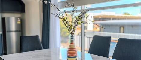 A modern dining area with a marble table, a colorful striped vase with branches, and a view of the city through large windows.