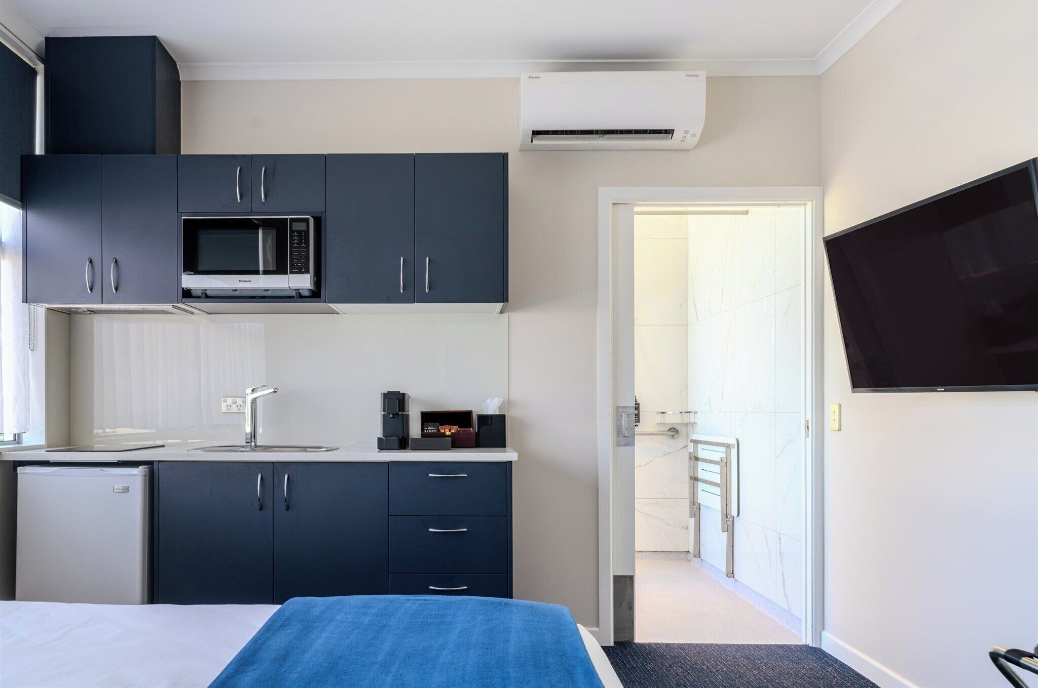 Habitación de hotel pequeña con kitchenette, una cama, un televisor y un baño a la vista a través de una puerta abierta.