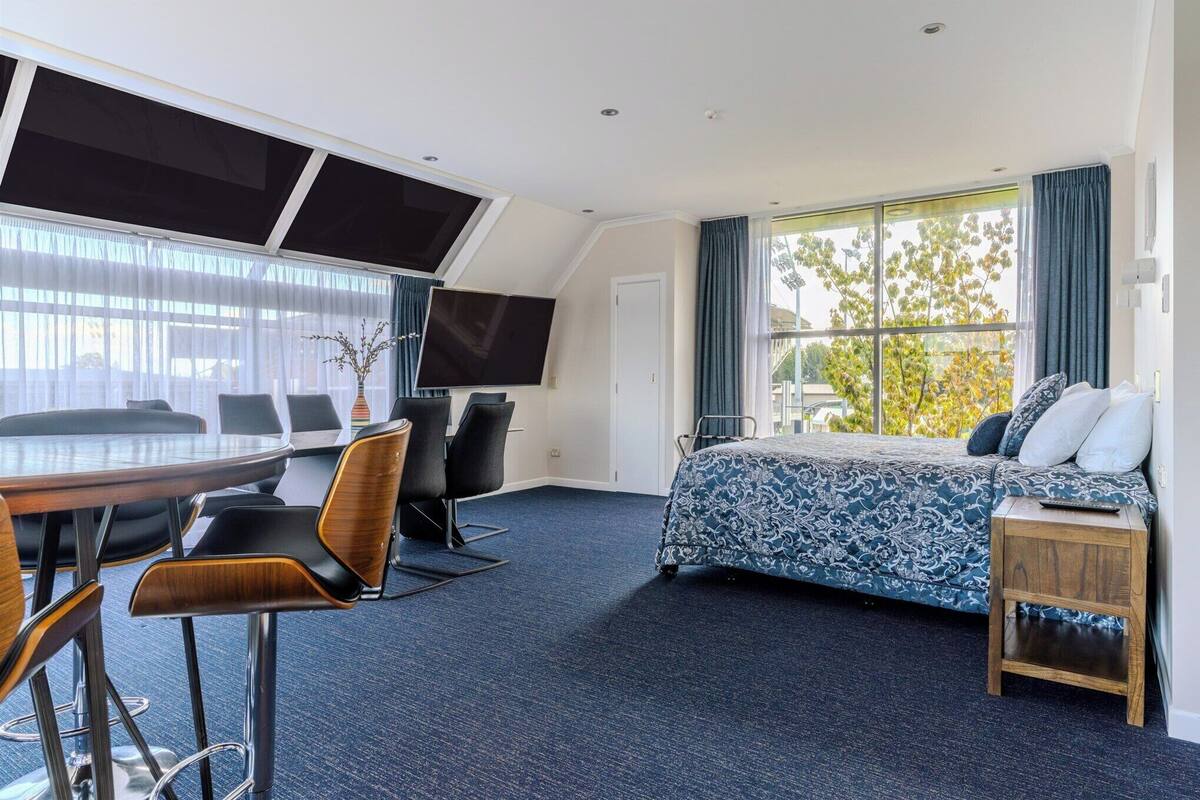 Une chambre d’hôtel avec un grand lit, un bureau avec des chaises, une télévision et une vue sur les arbres par la fenêtre.
