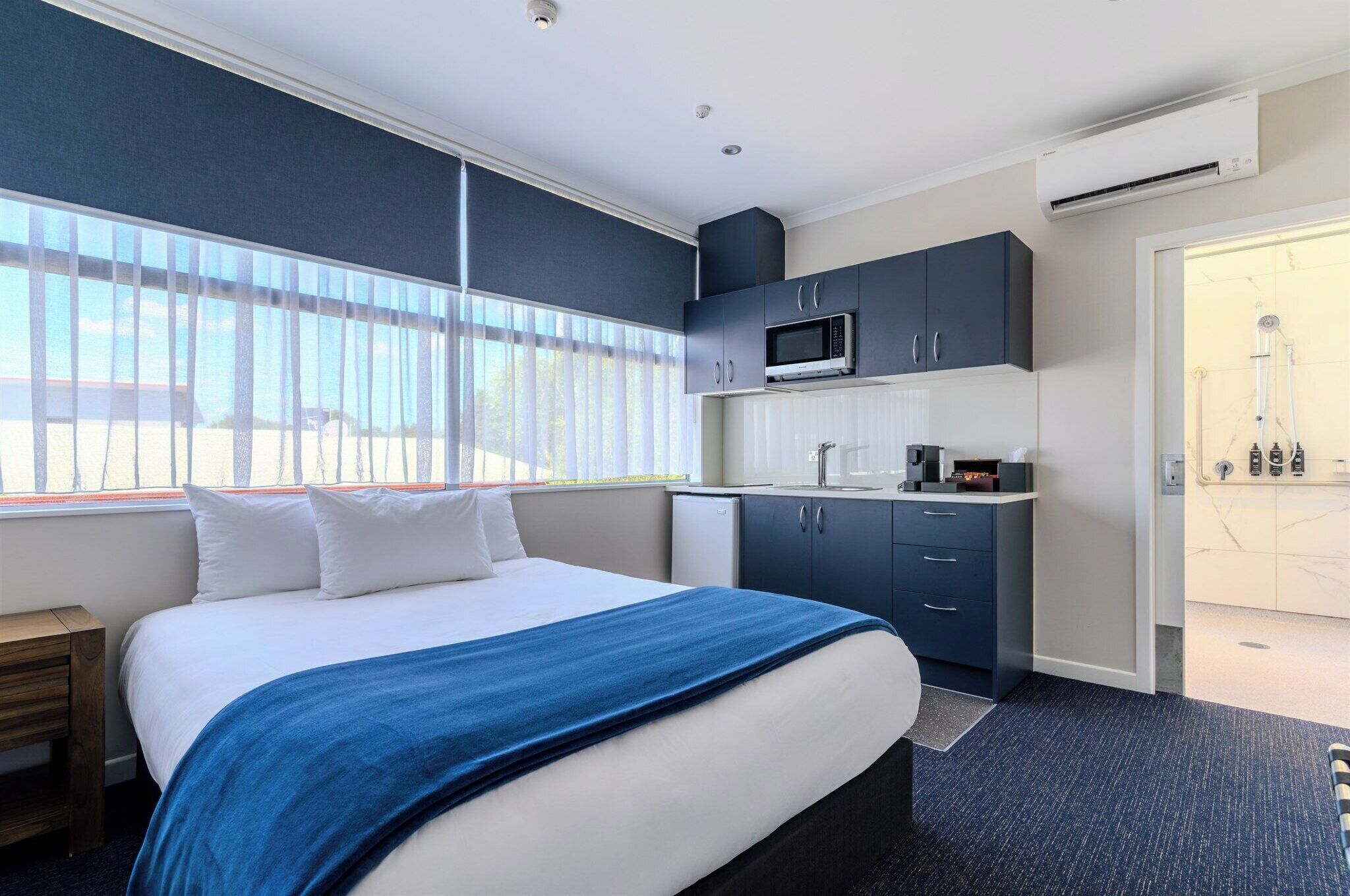 Habitación de hotel con cama, kitchenette pequeña, zona de ducha y ventana con persianas.